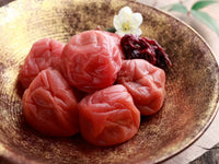 Close-up of pickled Ume (Prunus mume) fruits displayed in a decorative bowl with a single white flower garnish